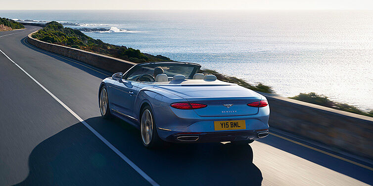 Bentley Padova New Bentley Continental GTC Azure convertible rear view in Light Windsor blue paint with 22 inch ten spoke wheels driving dynamically on an ocean road in the sunshine