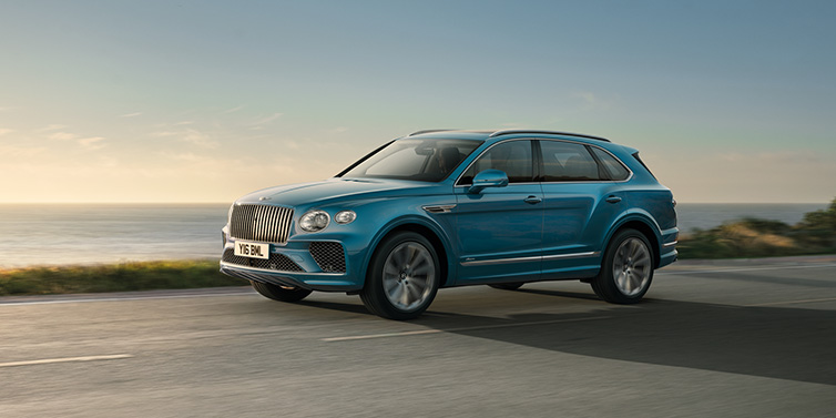 Bentley Padova Bentley Bentayga Azure SUV in Topaz blue paint driving dynamically by the ocean with 22 inch 10 spoke directional wheels