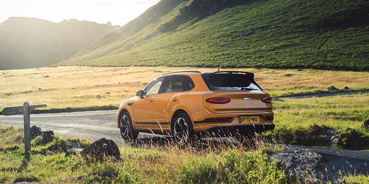 Bentley Padova Bentley Bentayga S SUV rear three quarter in Sunburst Gold paint on a road surrounded by green hills