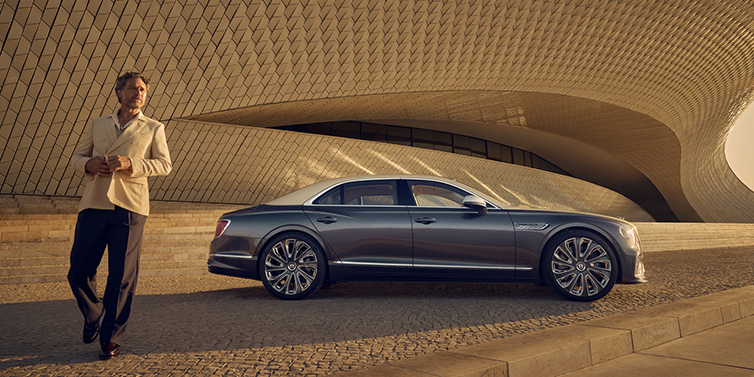 Bentley Padova Bentley Flying Spur Mulliner sedan in Rose Gold over Magnetic duotone paint parked by a curved wall with person in suit jacket standing on the left